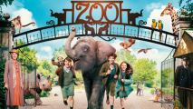 An elephant in the middle of three kids walking through a zoo entrance. There is a blue sky and green park in the background.